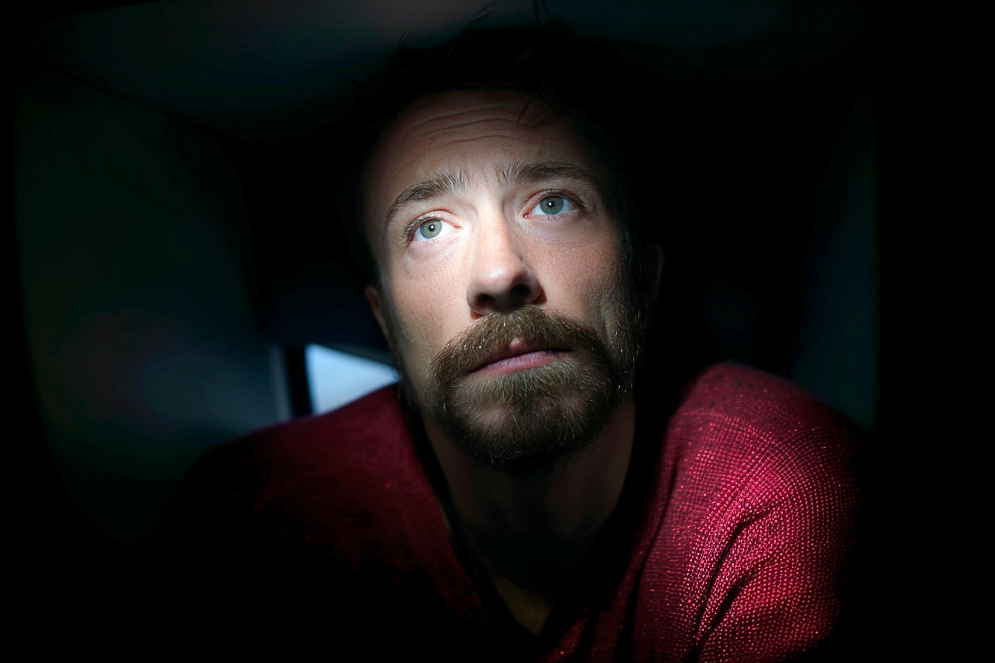 A man with a beard and mustache, wearing a red sweater, looks up into a beam of light in a dark room, with his face partially illuminated and an expression of contemplation or concern.