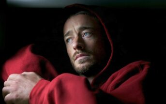 A man with light eyes and short facial hair wearing a red hooded sweatshirt, looking off to the side.