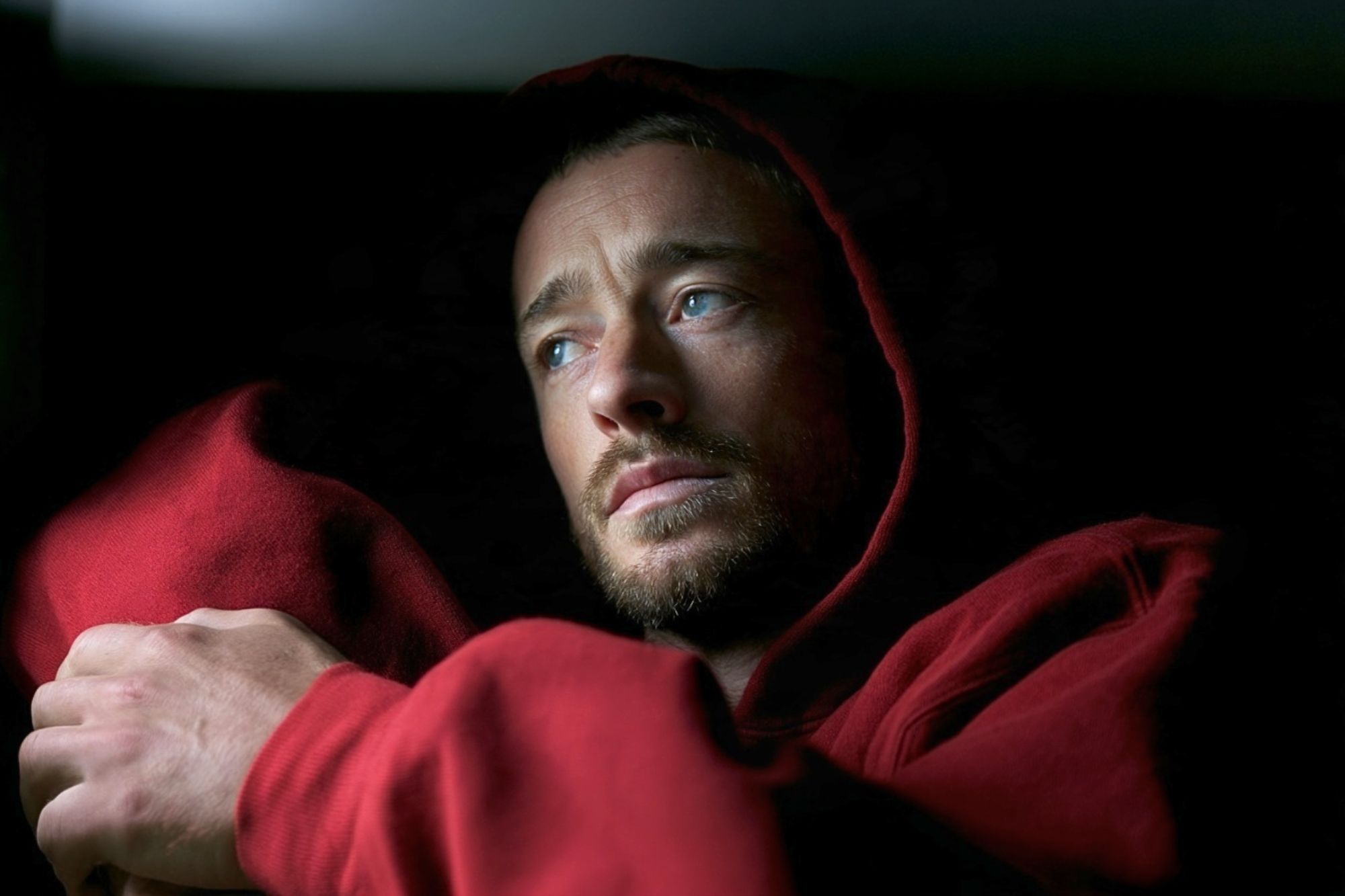 A man with light eyes and short facial hair wearing a red hooded sweatshirt, looking off to the side.