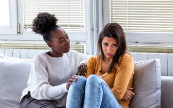 Two women sit on a couch indoors. One woman looks sad and hugs her knees, while the other gently touches her arm, offering comfort and support. They are surrounded by large windows with blinds.