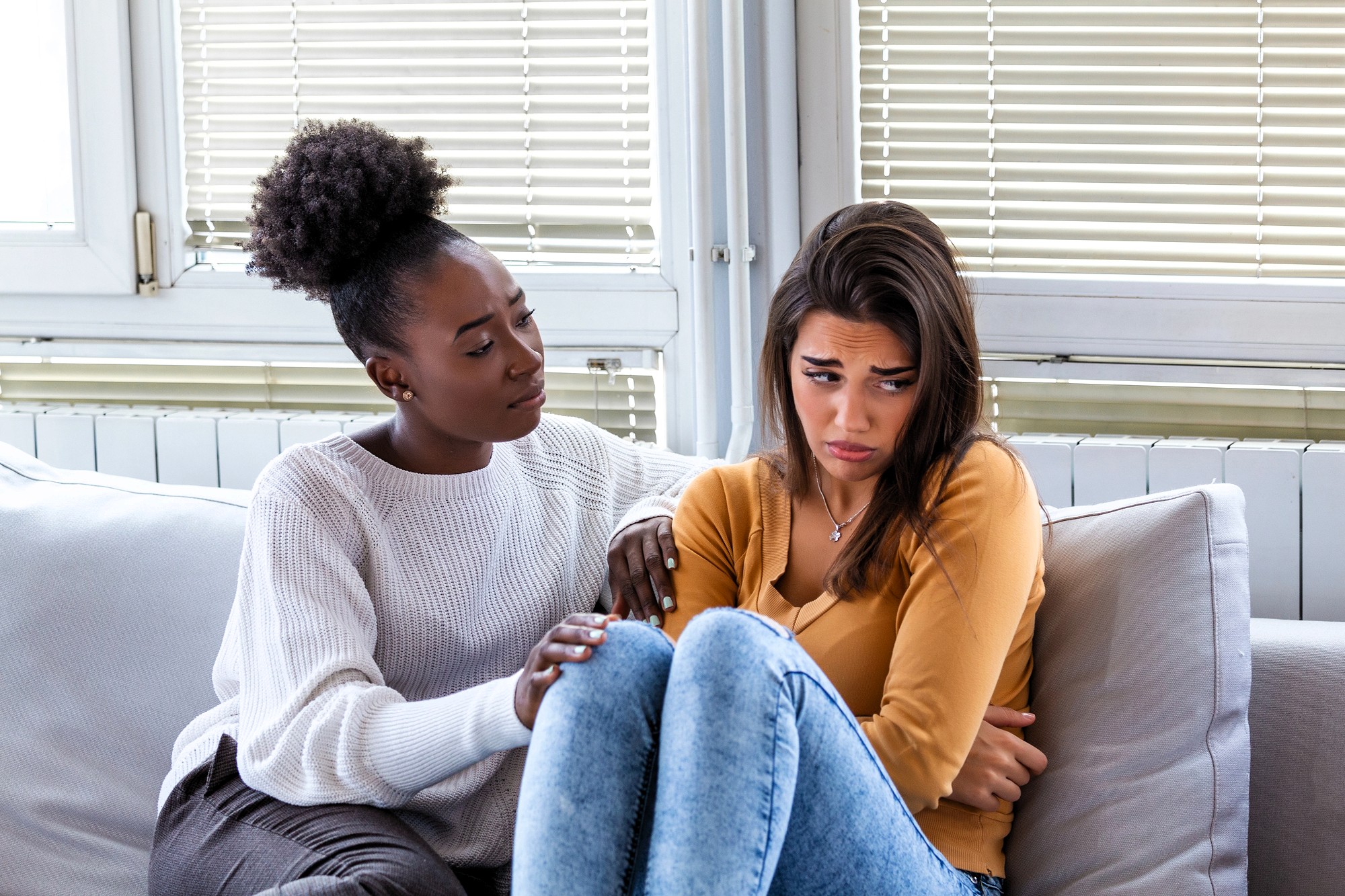 Two women sit on a couch indoors. One woman looks sad and hugs her knees, while the other gently touches her arm, offering comfort and support. They are surrounded by large windows with blinds.
