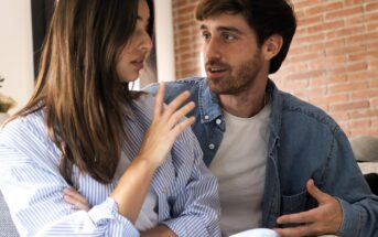 A woman and a man sit close together having a serious conversation. The woman looks away with her arms crossed while the man gestures with his hands, speaking earnestly. They are indoors with a brick wall behind them.