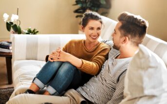 A woman and a man sit closely together on a light-colored sofa, smiling and looking at each other. The woman has her knees up and arms resting on them, while the man holds a mug. There are plants in the background.