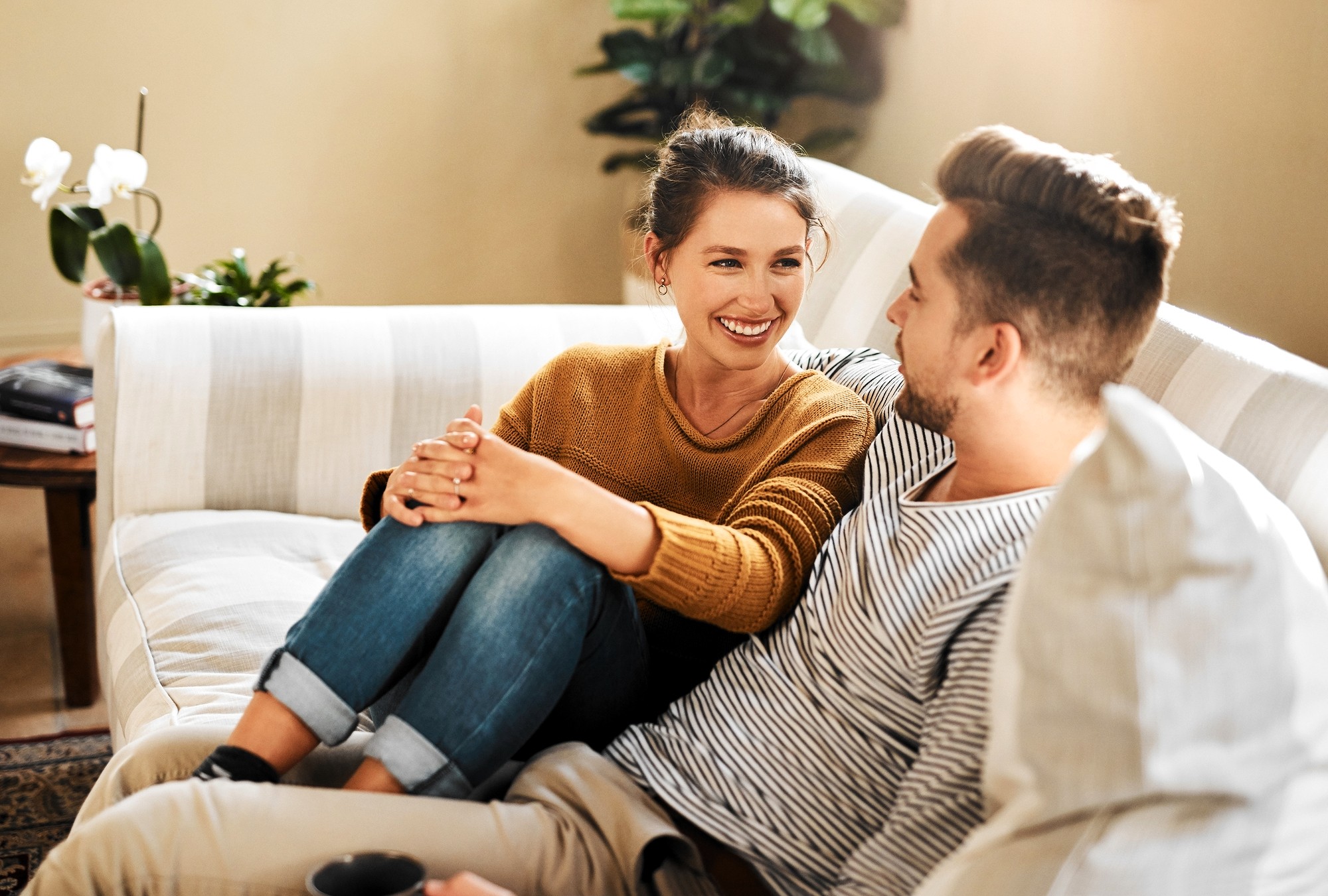 A woman and a man sit closely together on a light-colored sofa, smiling and looking at each other. The woman has her knees up and arms resting on them, while the man holds a mug. There are plants in the background.