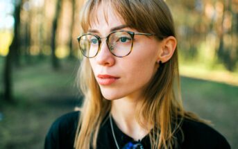 A young woman with light brown hair and glasses stands outdoors in a sunlit forest, looking thoughtfully into the distance. She wears a black top and a necklace. The background is softly blurred with trees.
