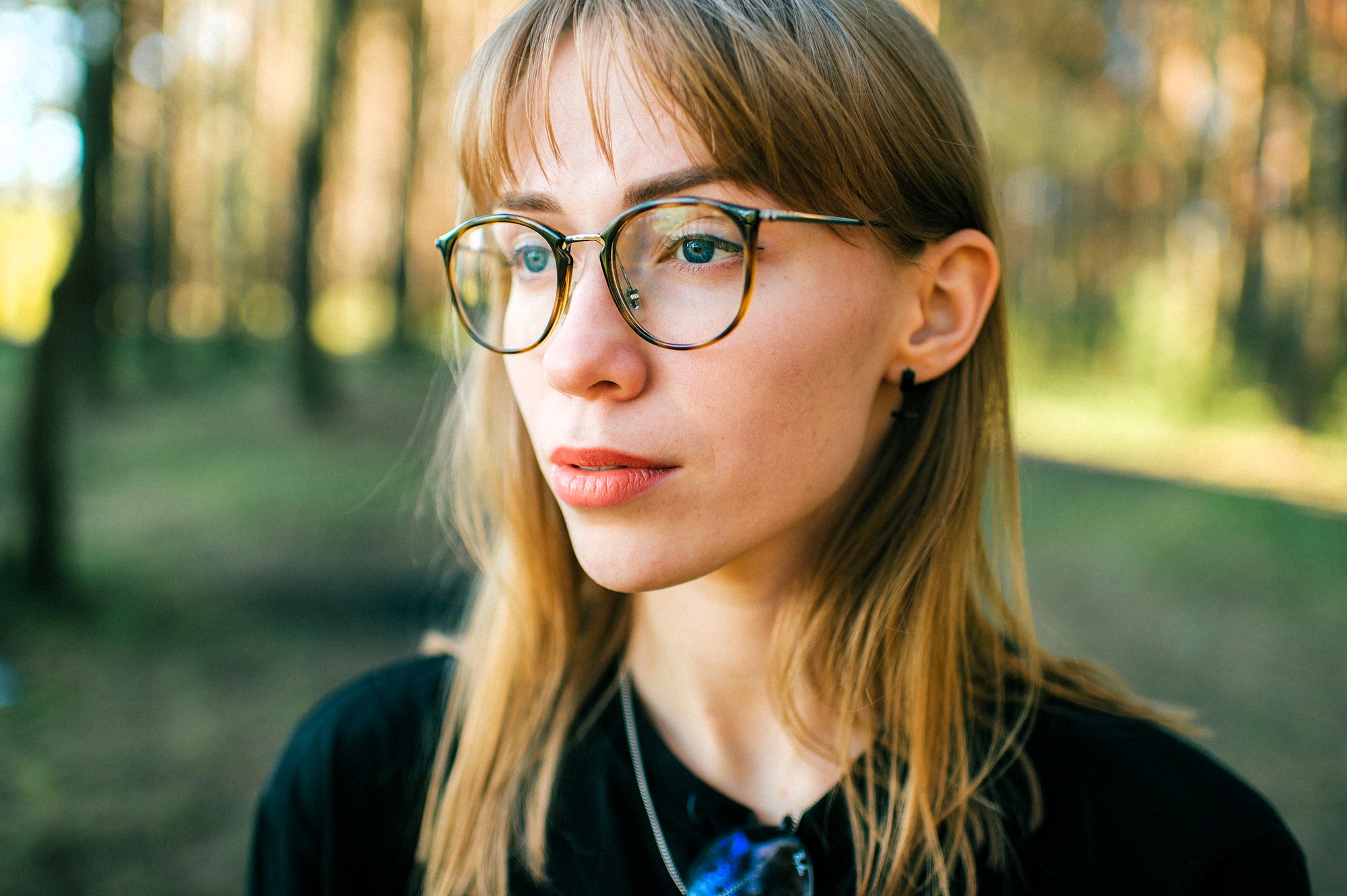 A young woman with light brown hair and glasses stands outdoors in a sunlit forest, looking thoughtfully into the distance. She wears a black top and a necklace. The background is softly blurred with trees.