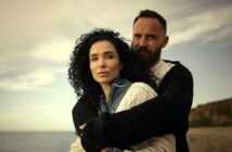 A man and woman stand on a beach, with the man embracing the woman from behind. Both have serious expressions. The woman has curly dark hair and wears a sweater; the man has a beard and wears a dark cardigan. The sky is cloudy.