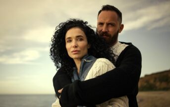 A man and woman stand on a beach, with the man embracing the woman from behind. Both have serious expressions. The woman has curly dark hair and wears a sweater; the man has a beard and wears a dark cardigan. The sky is cloudy.