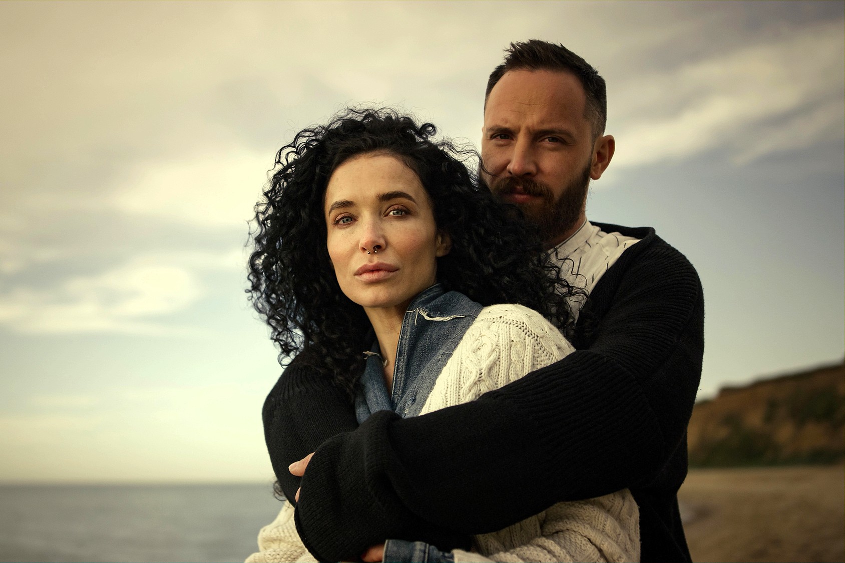 A man and woman stand on a beach, with the man embracing the woman from behind. Both have serious expressions. The woman has curly dark hair and wears a sweater; the man has a beard and wears a dark cardigan. The sky is cloudy.