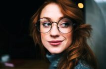 A person with long red hair, wearing round glasses and a gray turtleneck sweater, looks to the side and smiles softly against a dark background.