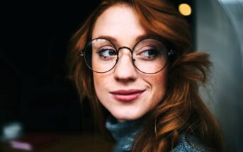 A person with long red hair, wearing round glasses and a gray turtleneck sweater, looks to the side and smiles softly against a dark background.