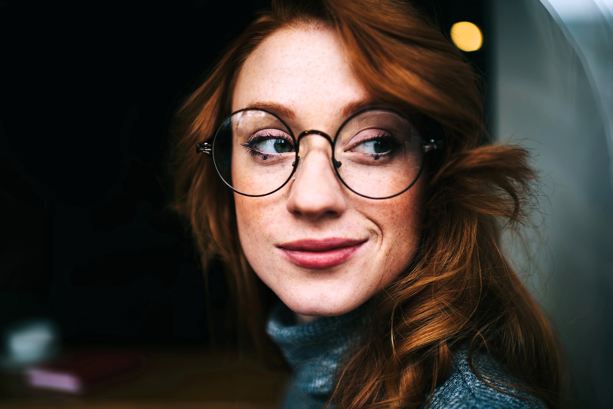 A person with long red hair, wearing round glasses and a gray turtleneck sweater, looks to the side and smiles softly against a dark background.