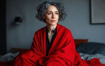 A woman with curly gray hair sits on a bed, wrapped in a bright red blanket, looking thoughtfully into the distance. The background is softly blurred in neutral tones.