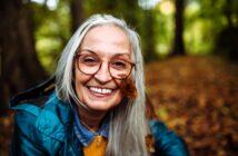 An older woman with long gray hair and glasses smiles outdoors in a forest. She wears a blue jacket, and a brown autumn leaf is caught in her hair and glasses. The background is filled with blurred trees and fallen leaves.