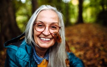 An older woman with long gray hair and glasses smiles outdoors in a forest. She wears a blue jacket, and a brown autumn leaf is caught in her hair and glasses. The background is filled with blurred trees and fallen leaves.