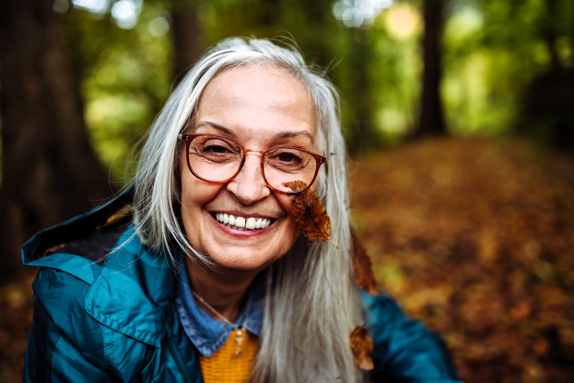 An older woman with long gray hair and glasses smiles outdoors in a forest. She wears a blue jacket, and a brown autumn leaf is caught in her hair and glasses. The background is filled with blurred trees and fallen leaves.