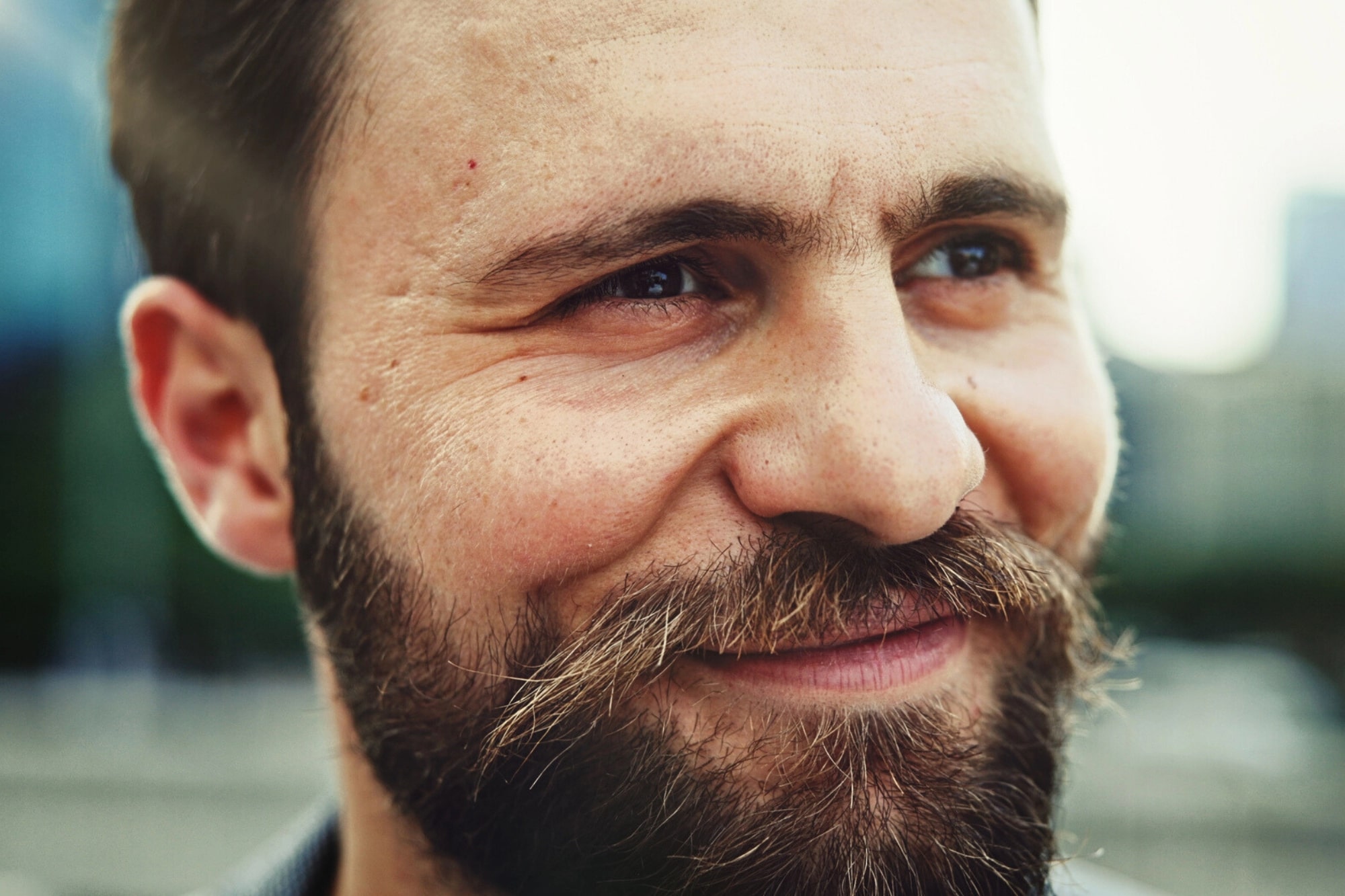 A close-up of a bearded man with brown hair, smiling softly and looking off to the side outdoors. The background is blurred, highlighting his facial features and expression.