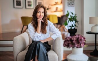 A woman with long brown hair, wearing a white blouse and black skirt, sits smiling in a modern, stylish living room with plants, flowers, and framed photos on the wall.