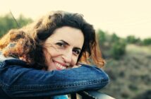A woman with curly brown hair and a denim jacket leans on a railing outdoors, smiling warmly at the camera with a blurred natural landscape in the background.
