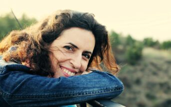 A woman with curly brown hair and a denim jacket leans on a railing outdoors, smiling warmly at the camera with a blurred natural landscape in the background.