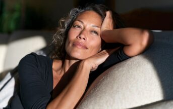 A woman with wavy hair resting her head on her hand while looking thoughtfully toward the light.