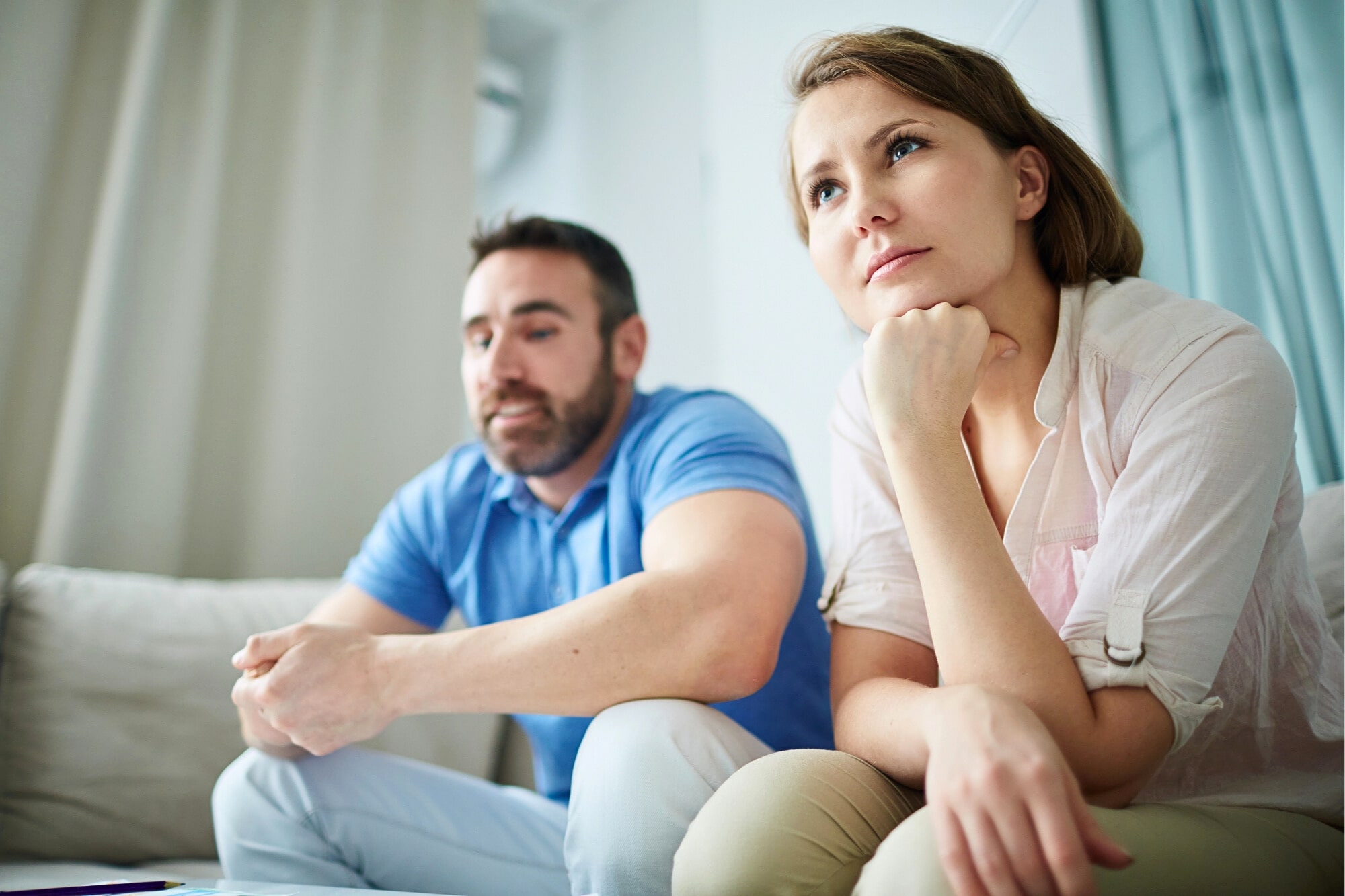 A woman and a man sit on a couch indoors, both looking serious and thoughtful. The woman is resting her chin on her hand, while the man sits with his hands clasped, gazing forward.