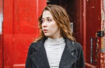 A young woman with wavy brown hair, wearing a gray coat and light turtleneck, looks to her left while standing in front of large, weathered red doors.