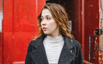 A young woman with wavy brown hair, wearing a gray coat and light turtleneck, looks to her left while standing in front of large, weathered red doors.