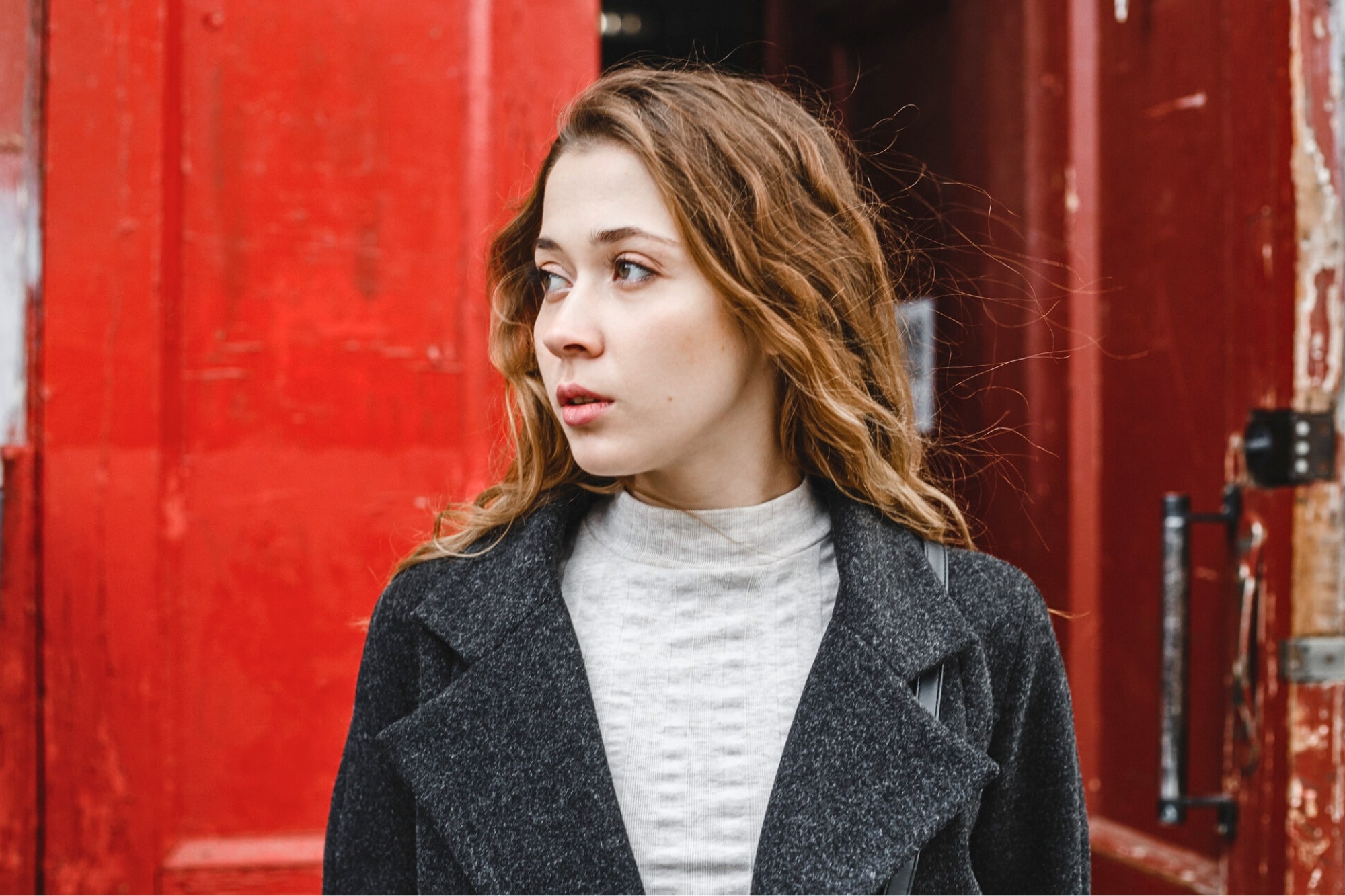 A young woman with wavy brown hair, wearing a gray coat and light turtleneck, looks to her left while standing in front of large, weathered red doors.
