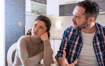 A woman sits at a table looking away with a bored or frustrated expression, resting her chin on her hand, while a man next to her gestures and speaks, appearing upset. They are in a modern kitchen.