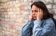 A woman in a denim jacket sits against a brick wall, eyes closed, holding her head with both hands, appearing stressed or in pain.