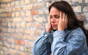 A woman in a denim jacket sits against a brick wall, eyes closed, holding her head with both hands, appearing stressed or in pain.