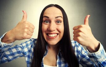 A smiling woman with long brown hair gives two thumbs up. She is wearing a blue and white plaid shirt and is standing in front of a plain gray background.