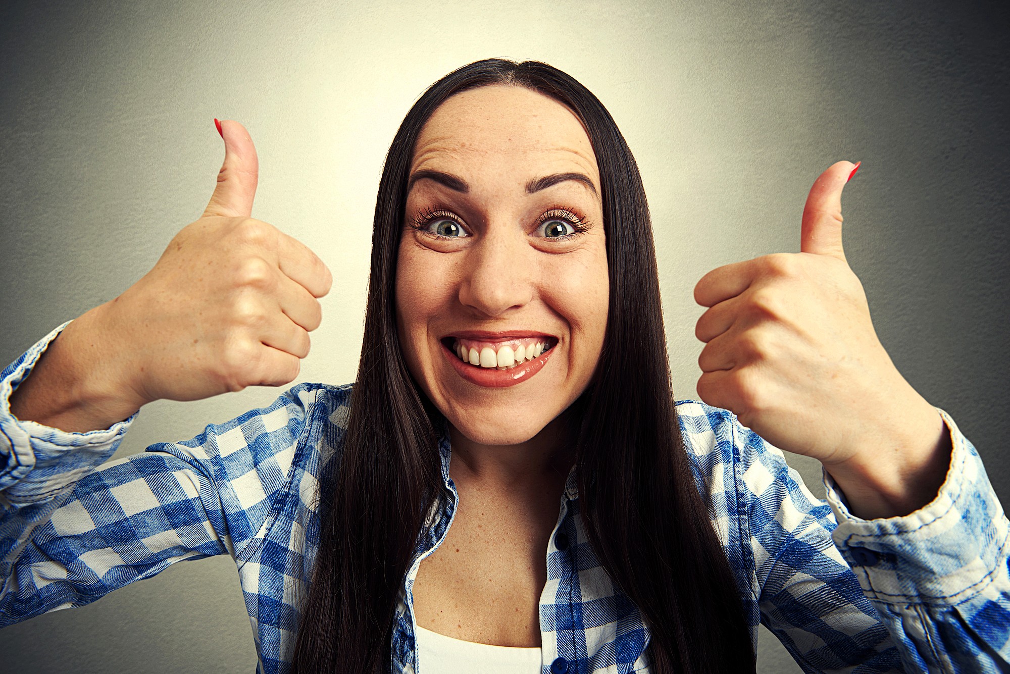 A smiling woman with long brown hair gives two thumbs up. She is wearing a blue and white plaid shirt and is standing in front of a plain gray background.