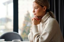 A woman with blonde hair in a ponytail, wearing a beige turtleneck sweater, sits at a table with her hands under her chin, gazing thoughtfully out of a large window.