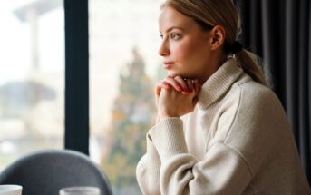 A woman with blonde hair in a ponytail, wearing a beige turtleneck sweater, sits at a table with her hands under her chin, gazing thoughtfully out of a large window.