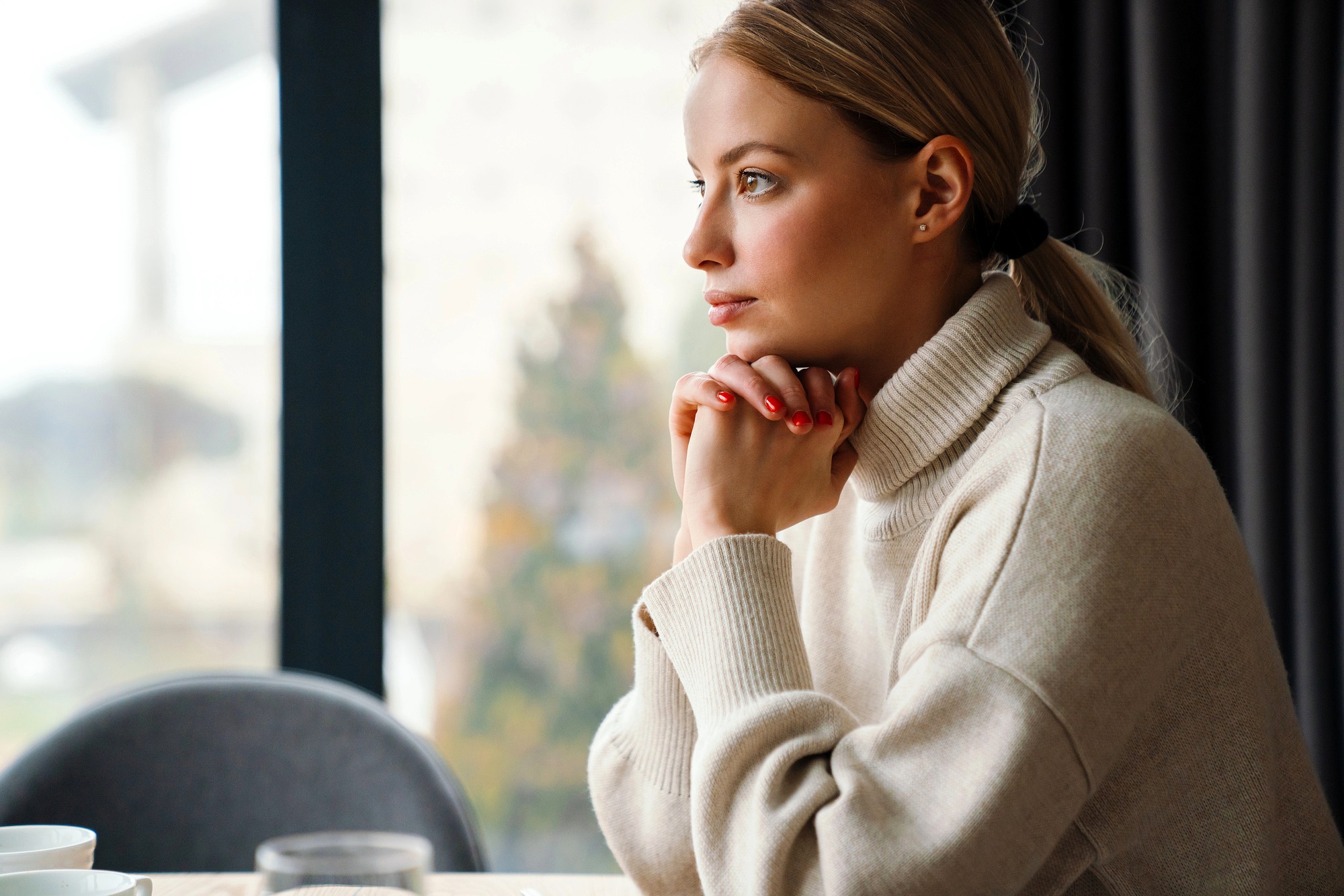 A woman with blonde hair in a ponytail, wearing a beige turtleneck sweater, sits at a table with her hands under her chin, gazing thoughtfully out of a large window.