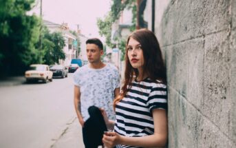 A young woman in a striped shirt leans against a stone wall, holding a hat, while a young man stands in the background on a quiet street lined with trees and parked cars.