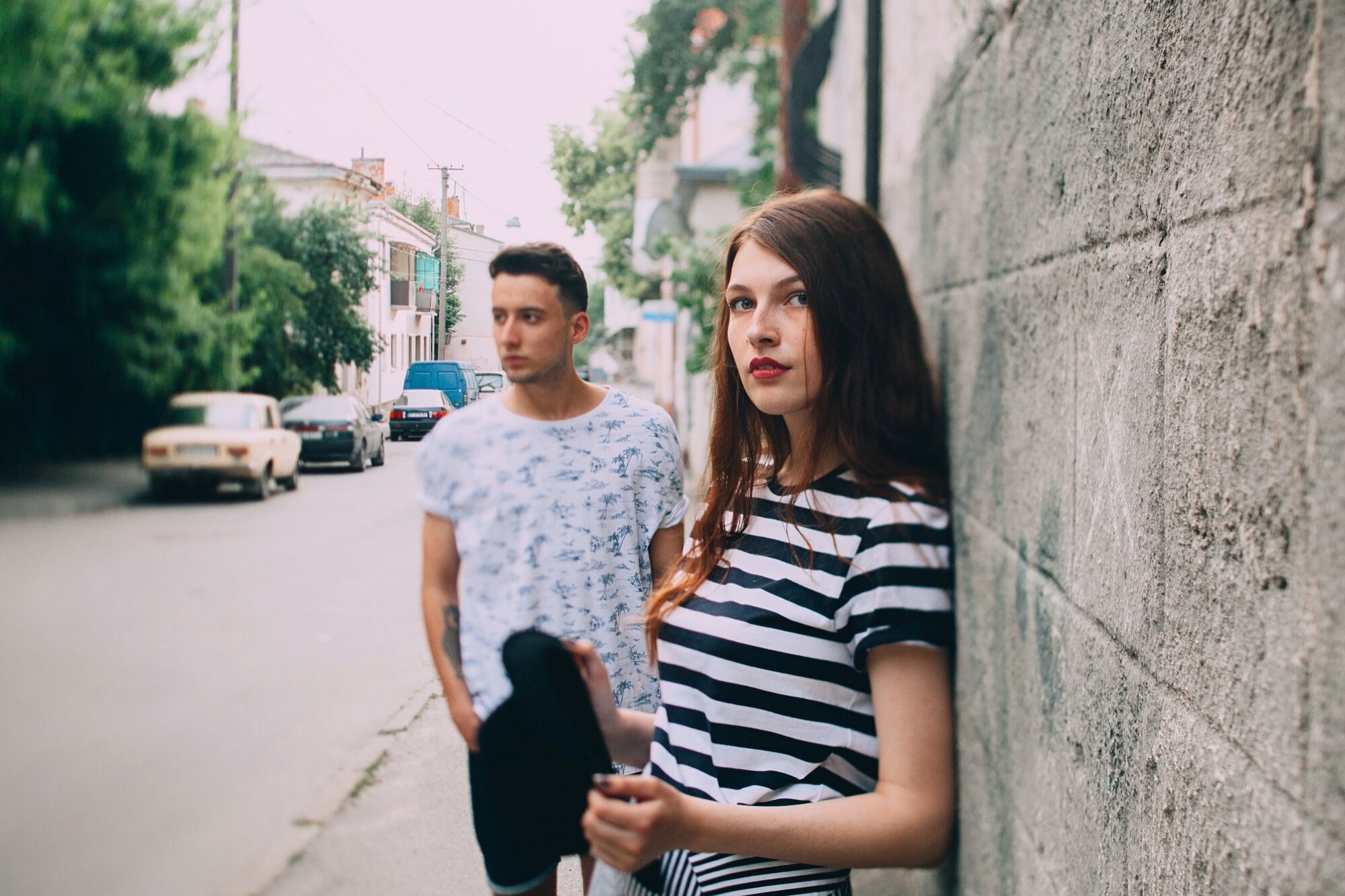 A young woman in a striped shirt leans against a stone wall, holding a hat, while a young man stands in the background on a quiet street lined with trees and parked cars.