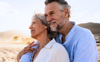 An older couple embraces and smiles while standing on a beach. Both wear light-colored clothing and look content, with sunlight highlighting their faces and mountains visible in the background.
