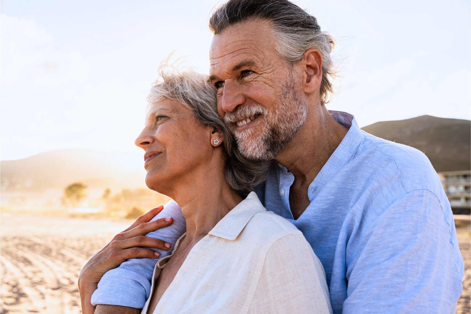 An older couple embraces and smiles while standing on a beach. Both wear light-colored clothing and look content, with sunlight highlighting their faces and mountains visible in the background.
