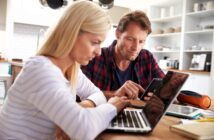 A woman and a man sit at a kitchen table, looking at a laptop and a smartphone. The woman types on the laptop while the man checks his phone, both appearing focused and engaged in their work.