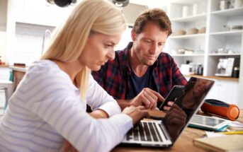 A woman and a man sit at a kitchen table, looking at a laptop and a smartphone. The woman types on the laptop while the man checks his phone, both appearing focused and engaged in their work.