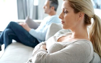 A woman with blonde hair in a ponytail sits on a couch looking thoughtful, with a man sitting in the background facing away, both appearing distant and quiet.
