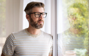 A man with glasses and a beard, wearing a beige and white striped t-shirt, stands indoors by a window, gazing outside with a thoughtful expression. His reflection is visible in the glass.