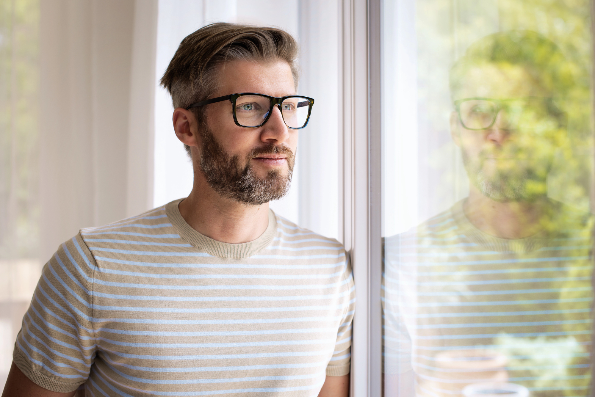A man with glasses and a beard, wearing a beige and white striped t-shirt, stands indoors by a window, gazing outside with a thoughtful expression. His reflection is visible in the glass.