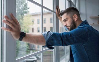 A man wearing a denim shirt and smartwatch stands indoors, leaning against large window panes with both hands, looking outside thoughtfully on a cloudy day.