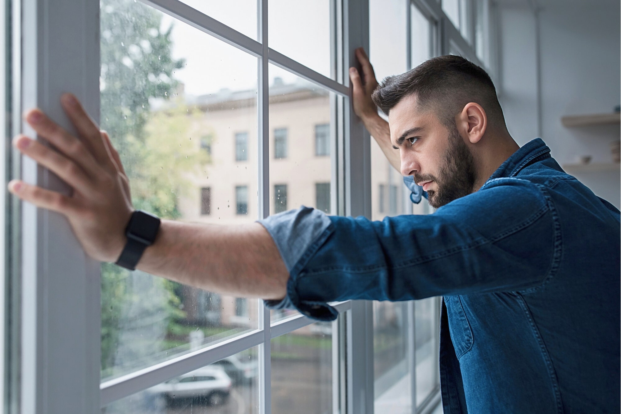A man wearing a denim shirt and smartwatch stands indoors, leaning against large window panes with both hands, looking outside thoughtfully on a cloudy day.