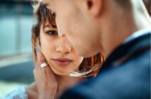 A close-up of a woman with brown hair looking into the camera while a man gently holds her face, his back partially to the camera. Soft natural light highlights their intimate moment.