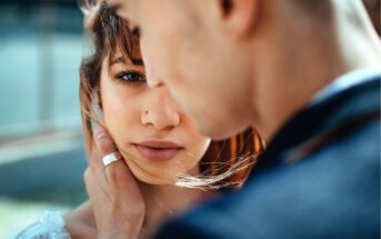 A close-up of a woman with brown hair looking into the camera while a man gently holds her face, his back partially to the camera. Soft natural light highlights their intimate moment.